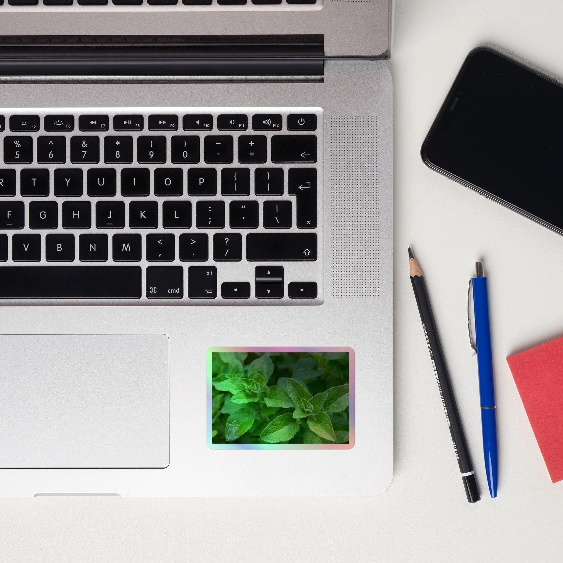 Lifestyle photo of a rectangular holographic sticker with a close-up image of vivid green oregano leaves, placed on a laptop to the right of the mouse pad.