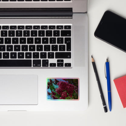 Lifestyle photo of a rectangular holographic sticker with an image of bright pink crape myrtle blooms with green leaves and blue sky in the background, placed on a laptop to the right of the mouse pad.