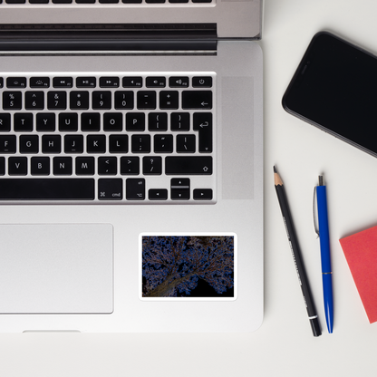 Lifestyle photo of a rectangular vinyl sticker with an image of a neon blue and yellow tree design with lightning like features, placed on a laptop to the right of the mouse pad.