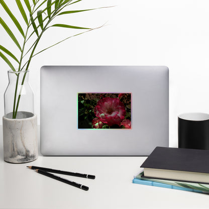 Lifestyle photo of a rectangular holographic sticker with an image of bright pink rose blooms with white centers, placed on a laptop with desk supplies nearby.