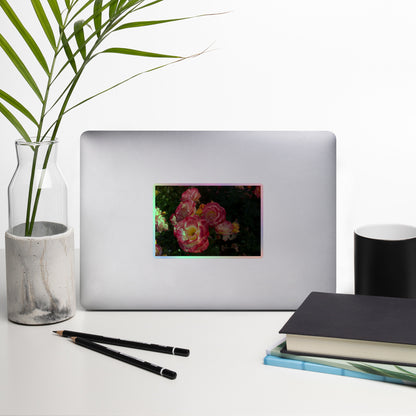 Lifestyle photo of a rectangular holographic sticker with an image of pink, yellow and white rose blooms with dark green leaves, placed on a laptop with desk supplies nearby.