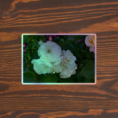Lifestyle photo of a rectangular holographic sticker with an image of a cluster of white roses with dark green leaves, placed on a brown wooden table surface.