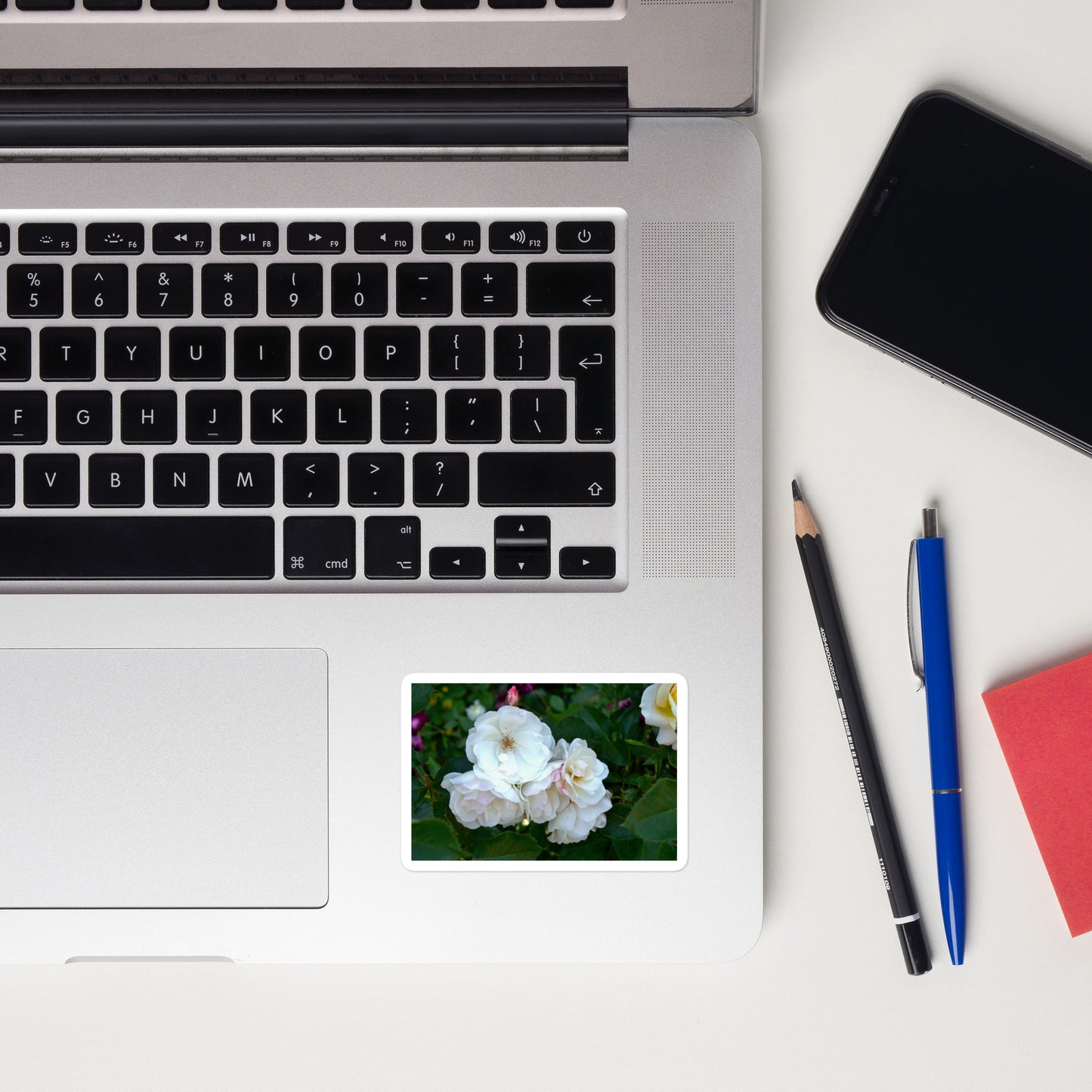 Lifestyle photo of a rectangular vinyl sticker with an image of white roses among dark green leaves, placed on a laptop to the right of the mouse pad.