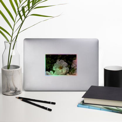 Lifestyle photo of a rectangular holographic sticker with an image of a white rose bloom with a honeybee on it, placed on a laptop with desk supplies nearby.