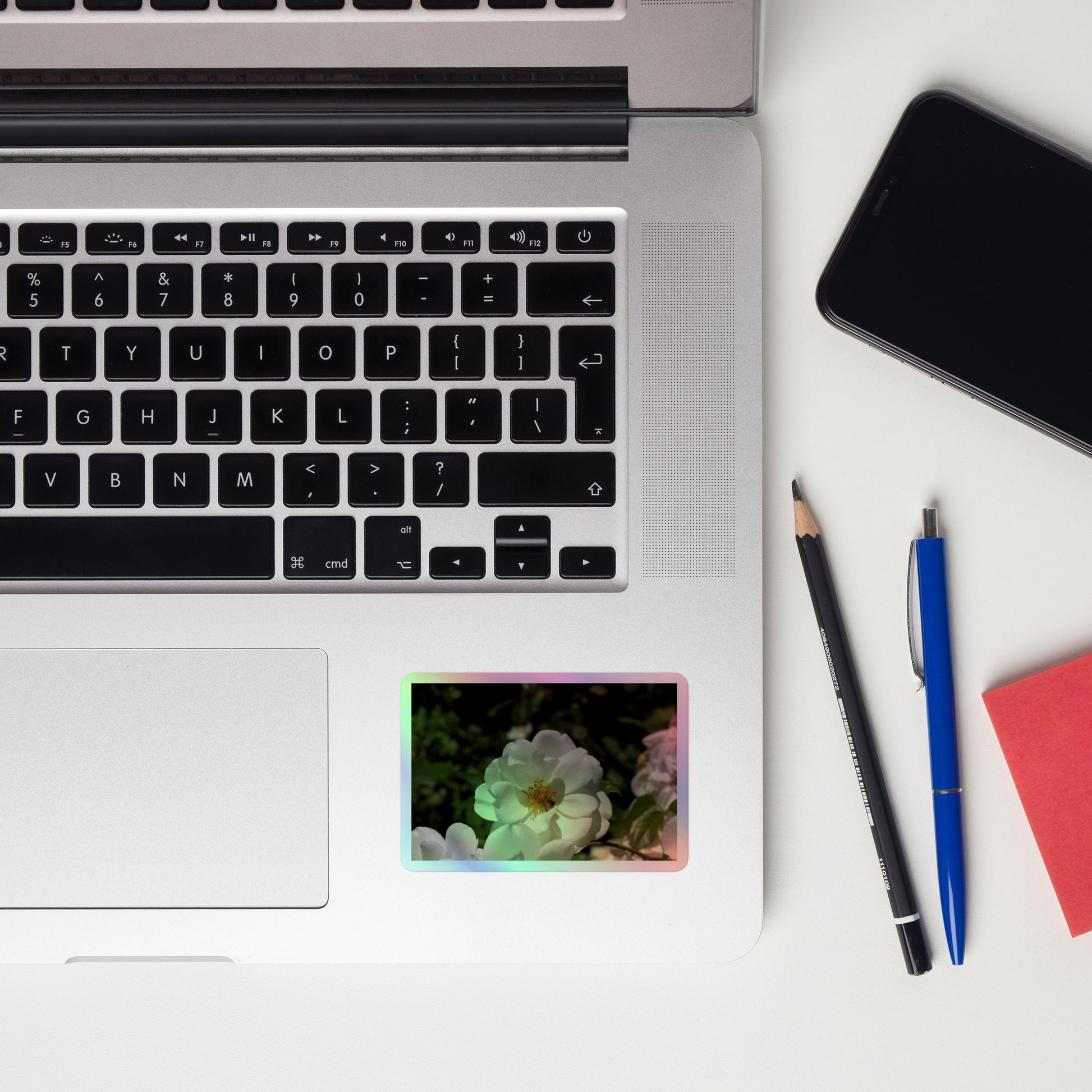 Lifestyle photo of a rectangular holographic sticker with an image of a white rose bloom with a honeybee on it, placed on a laptop to the right of the mouse pad.