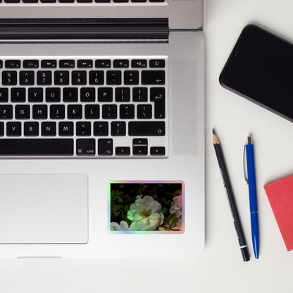 Lifestyle photo of a rectangular holographic sticker with an image of a white rose bloom with a honeybee on it, placed on a laptop to the right of the mouse pad.