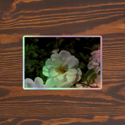 Lifestyle photo of a rectangular holographic sticker with an image of a white rose bloom with a honeybee on it, placed on a brown wooden table surface.