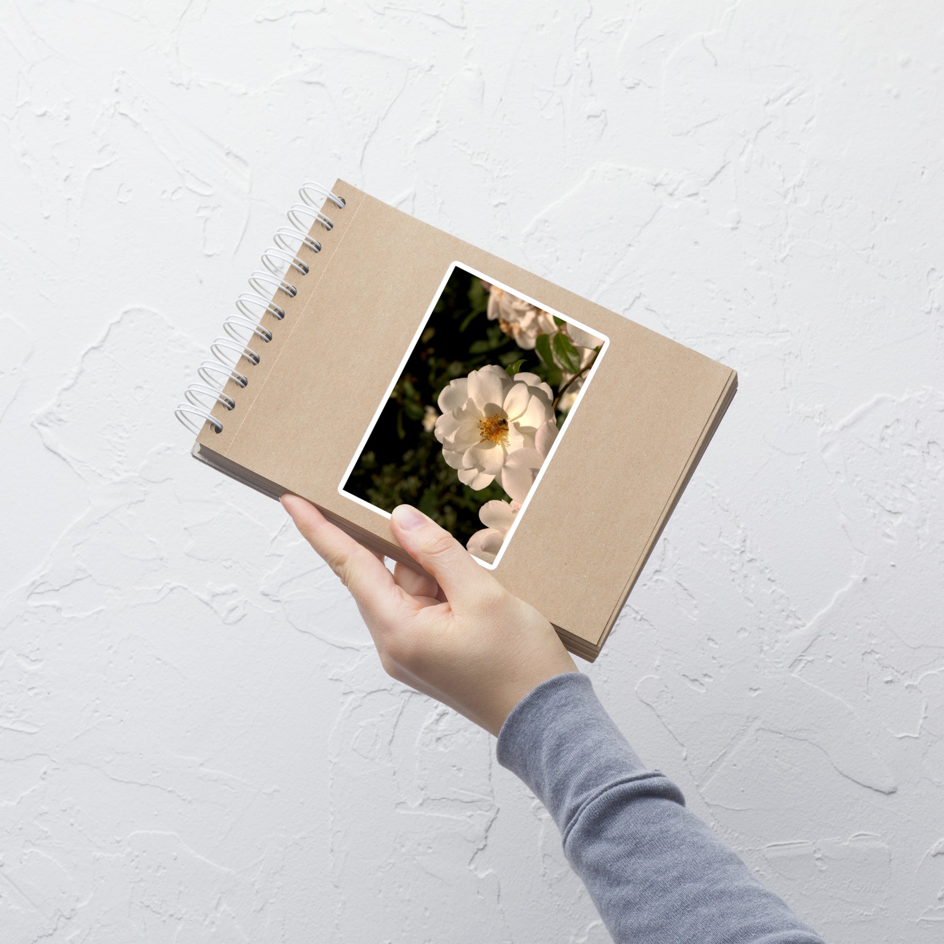Lifestyle photo of a rectangular vinyl sticker with an image of a white rose with a honeybee on it, placed on a small notepad.