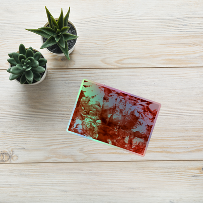 Lifestyle photo of a rectangular holographic sticker with an image of a vivid red water reflection design, placed on a table with two small succulents.