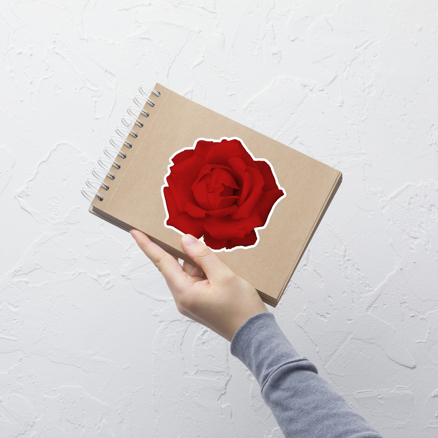 Lifestyle photo of a kiss-cut vinyl sticker with an image of a vivid red rose design, placed on a small notepad.