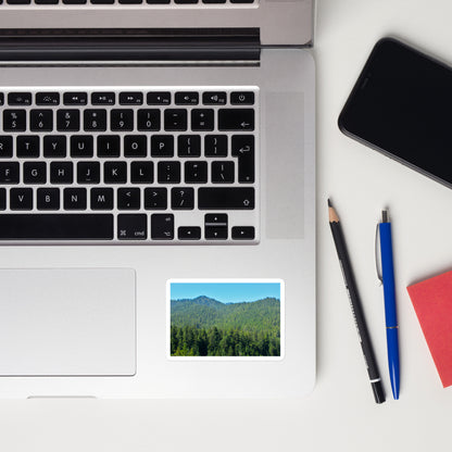 Lifestyle photo of a rectangular vinyl sticker with an image of mountain landscape with redwood trees and blue sky, placed on a laptop to the right of the mouse pad.
