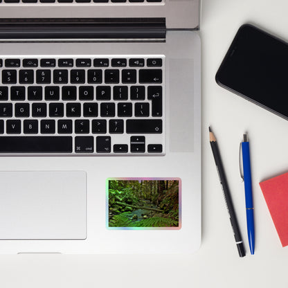 Lifestyle photo of a rectangular holographic sticker with an image of a creek through a redwood forest with ferns, placed on a laptop with desk supplies nearby.