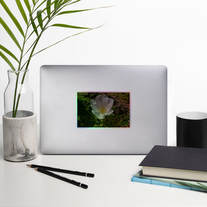 Lifestyle photo of a rectangular holographic sticker with a close-up image of a white flower with yellow stamen, placed on a laptop with desk supplies nearby.