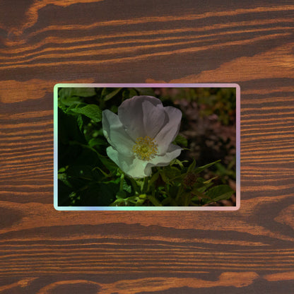 Lifestyle photo of a rectangular holographic sticker with a close-up image of a white flower with yellow stamen, placed on a brown wooden table surface.