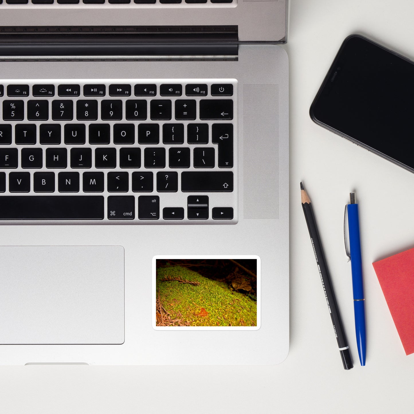 Lifestyle photo of a rectangular vinyl sticker with an image of green moss on a fallen tree, placed on a laptop to the right of the mouse pad.