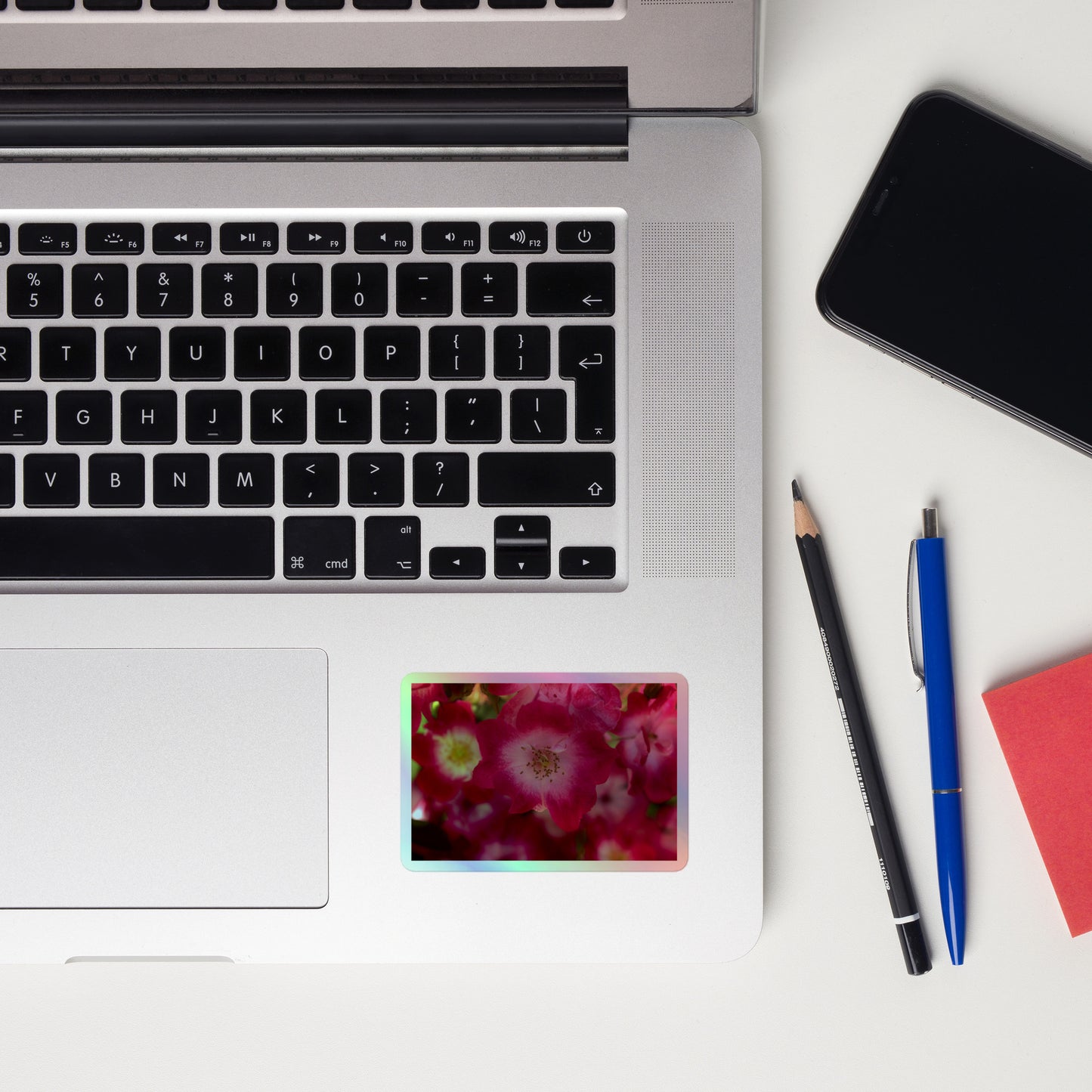 Lifestyle photo of a rectangular holographic sticker with an image of pink rose blooms with white centers, placed on a laptop to the right of the mouse pad.