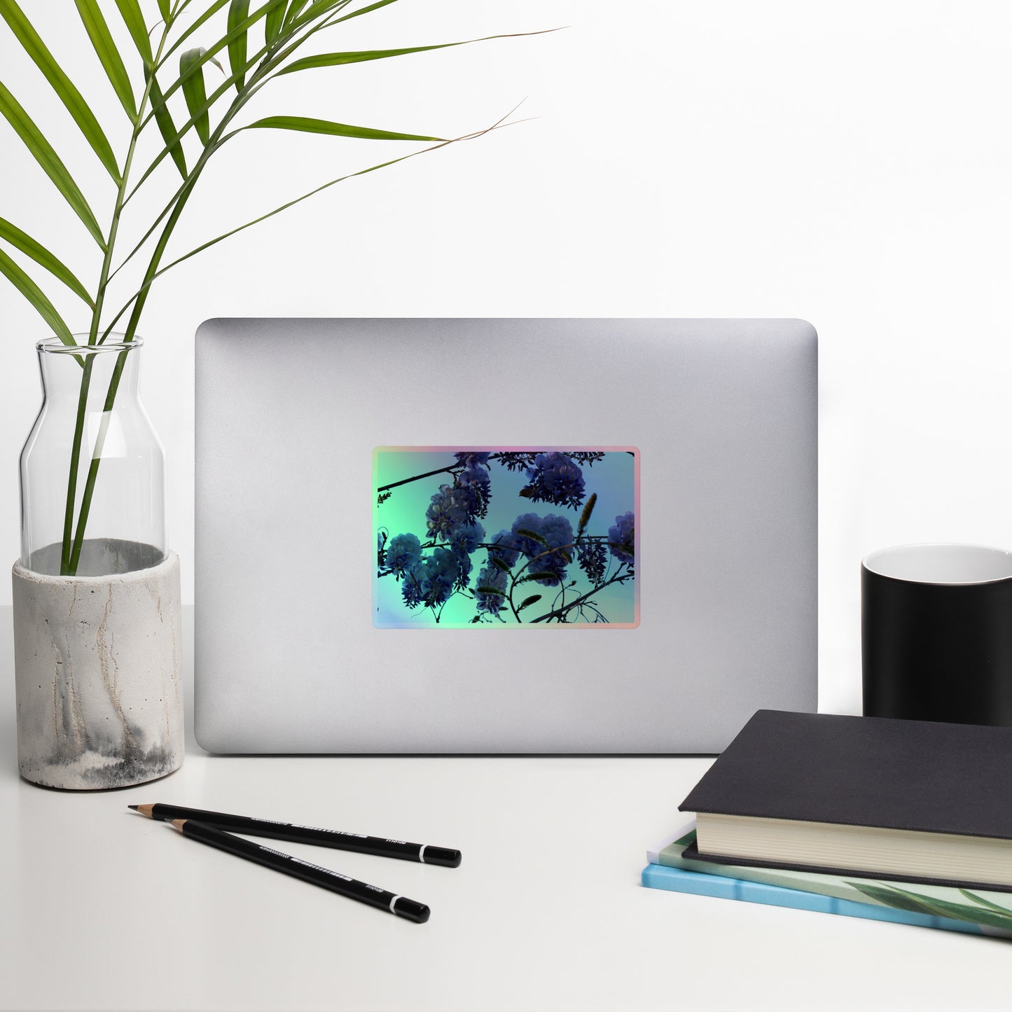 Lifestyle photo of a rectangular holographic sticker with an image of purple wisteria blooms with blue sky in the background, placed on a laptop with desk supplies nearby.