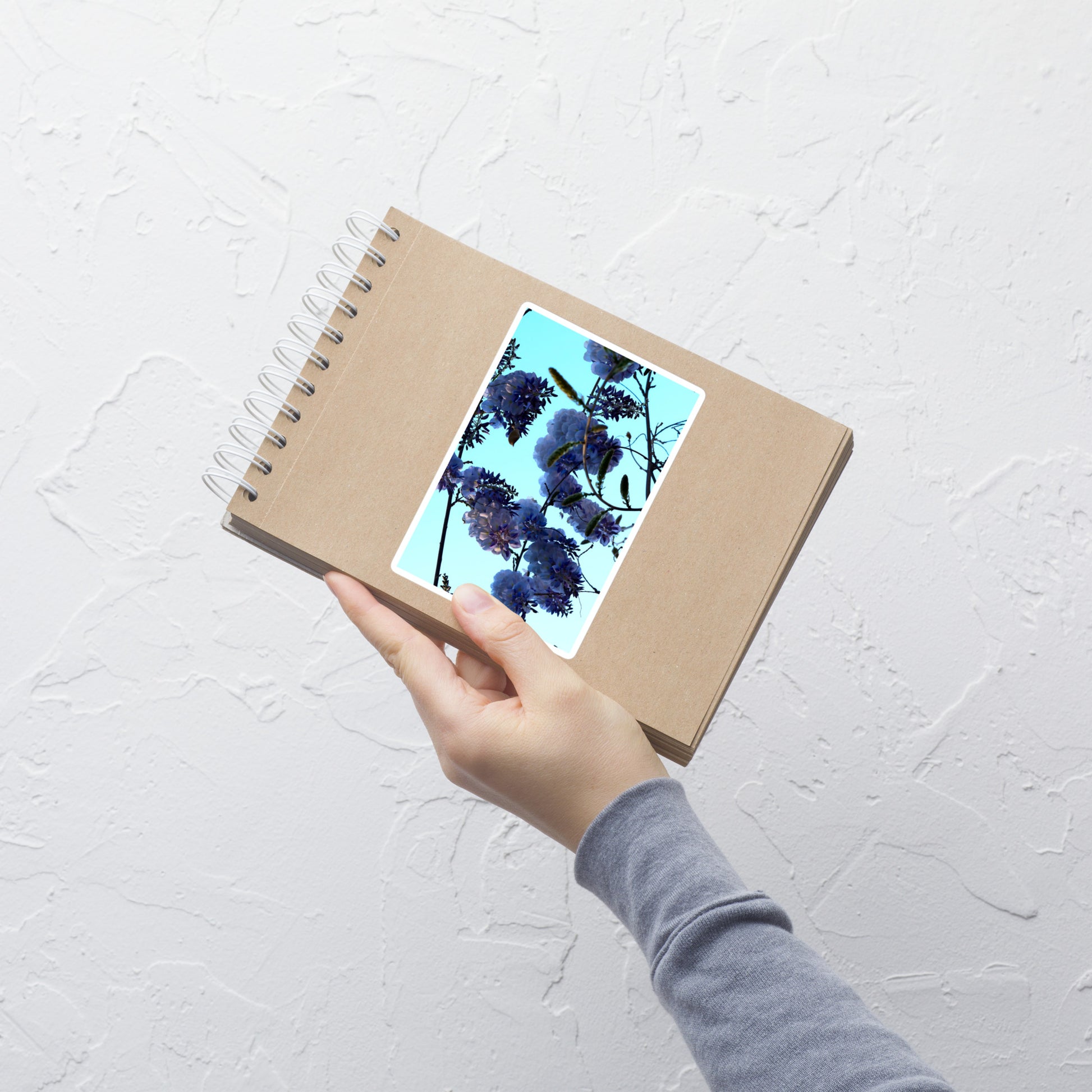 Lifestyle photo of a rectangular vinyl sticker with an image of a blue sky and purple wisteria blooms, placed on a small notepad.