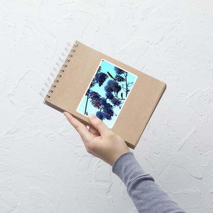 Lifestyle photo of a rectangular vinyl sticker with an image of a blue sky and purple wisteria blooms, placed on a small notepad.
