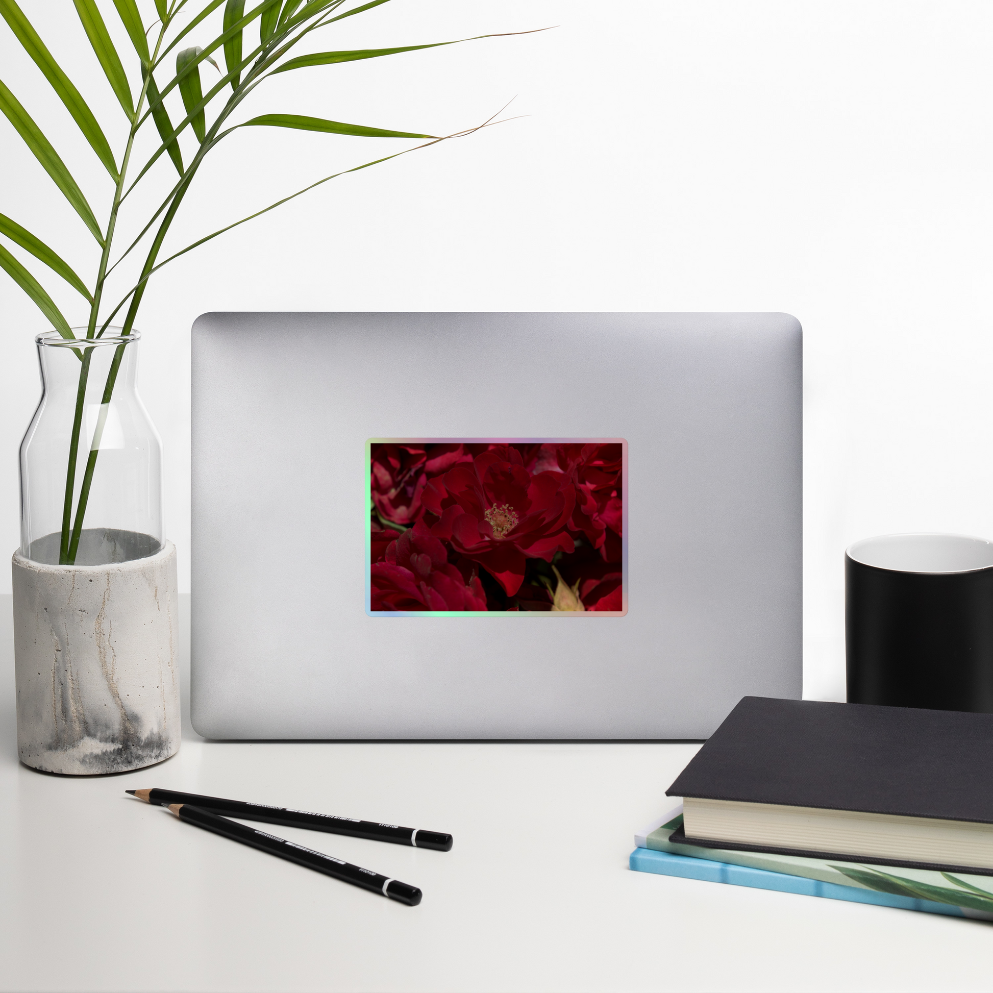Lifestyle photo of a rectangular holographic sticker with a close-up image of dark red roses, placed on a laptop with desk supplies nearby.