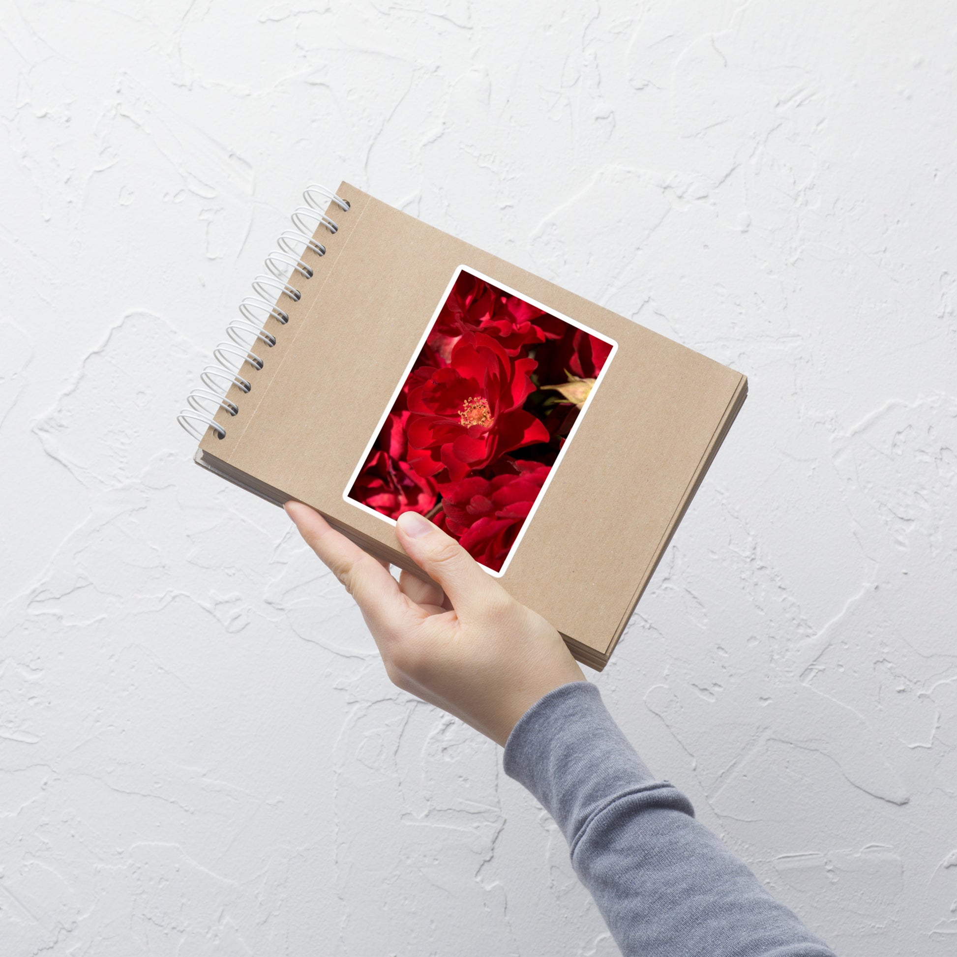 Lifestyle photo of a rectangular vinyl sticker with an image of bright red rose blooms with visible stamen, placed on a small notepad.