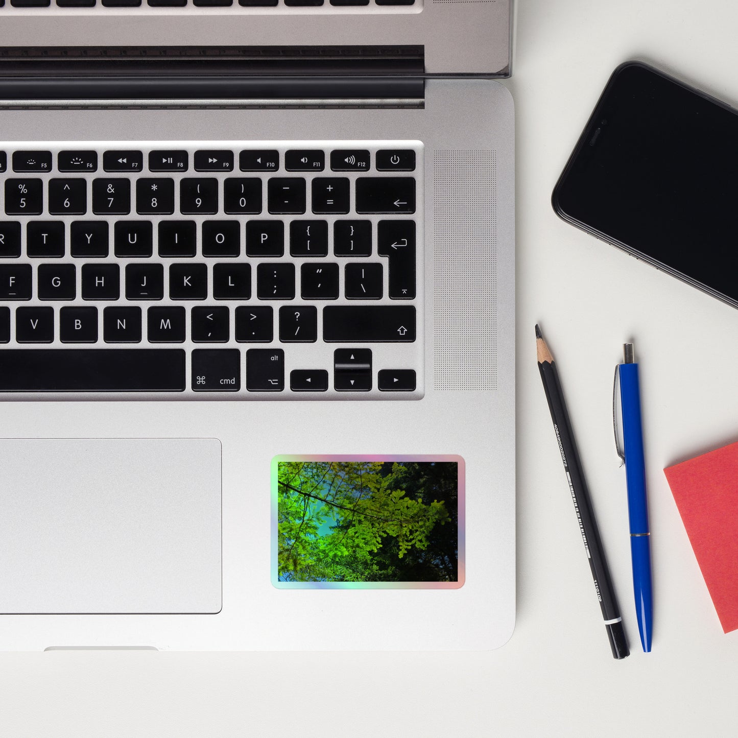 Lifestyle photo of a rectangular holographic sticker with an image of a redwood tree branch with blue sky behind it, placed on a laptop to the right of the mouse pad.