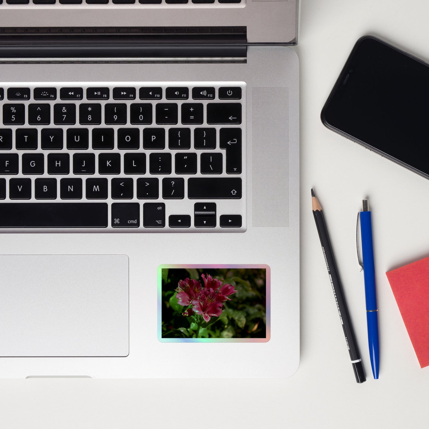 Lifestyle photo of a rectangular holographic sticker with a close-up image of vivid pink Peruvian Lilies with yellow accents, placed on a laptop to the right of the mouse pad.