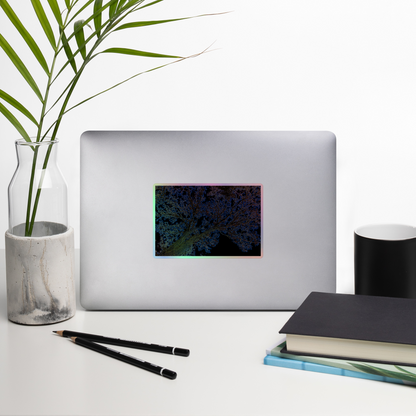 Lifestyle photo of a rectangular holographic sticker with an image of a yellow and blue neon tree lightning design, placed on a laptop with desk supplies nearby.