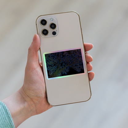 Lifestyle photo of a rectangular holographic sticker with an image of a yellow and blue neon tree lightning design, placed on the back of a phone.